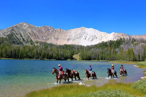 People ride horses through the shallow water of a lake in front of a mountain.