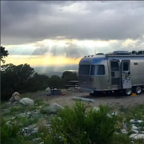 A silver travel trailer is parked at a scenic campsite beneath cloudy skies.