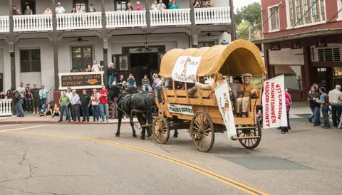 A covered wagon pulled by horses drives down a street during an event.