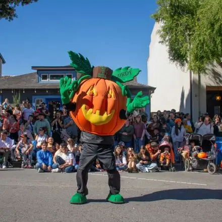 A person in a pumpkin mascot costume performs in front of a crowd on a street.