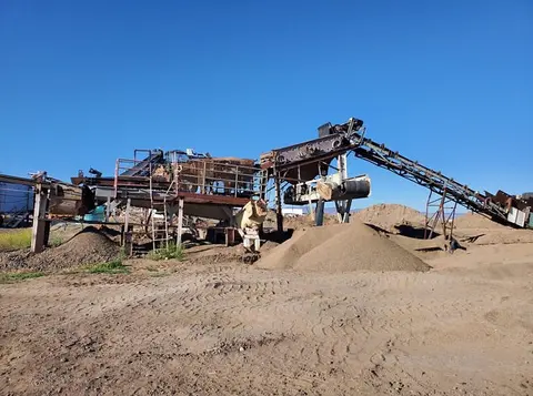 An industrial rock crusher and conveyor system stands in a dirt lot under a clear blue sky.