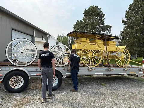 Two people load restored horse-drawn carriages onto a flatbed trailer.