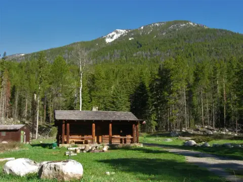 A log cabin sits in a clearing surrounded by a forest with a mountain in the background.