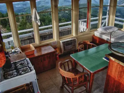 The cozy interior of a fire lookout tower with a stove, table, and bed.