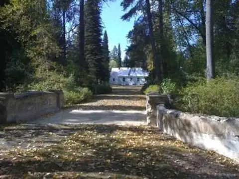 A stone bridge leads down a path towards a white building surrounded by trees.