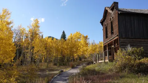 An old wooden building stands beside a dirt path surrounded by trees with yellow autumn leaves under a blue sky.