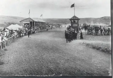 A historic black and white photo shows a crowd gathered at a dirt race track with vehicles parked nearby.
