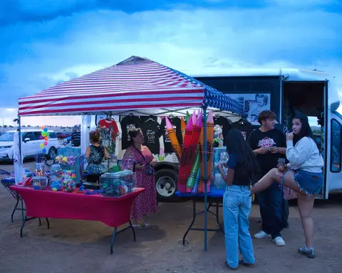 People gather at a vendor booth under an American flag tent next to a vehicle outdoors.