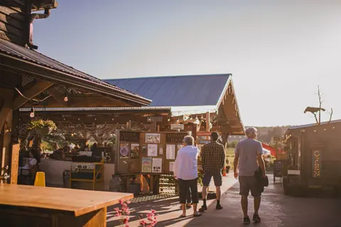 People walking past rustic buildings and outdoor information boards on a sunny day.