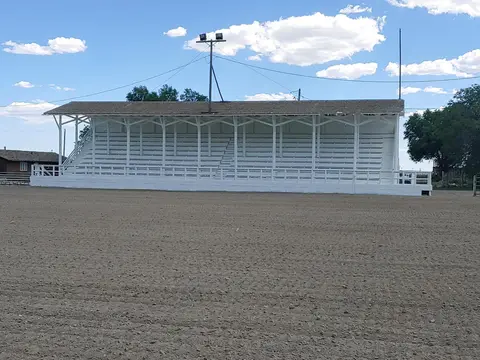 A white wooden grandstand stands by a dirt arena under a cloudy blue sky.