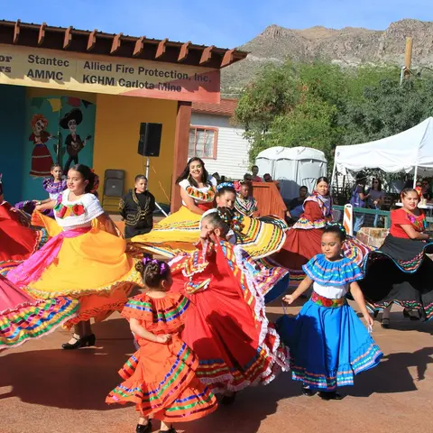 A group of girls in colorful traditional dresses perform a folk dance outdoors.