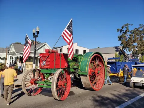 A vintage tractor decorated with American flags is displayed on a street with people nearby.