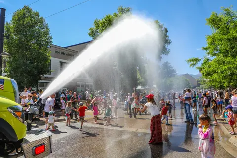 A crowd of people enjoys water being sprayed from a fire truck on a sunny street.