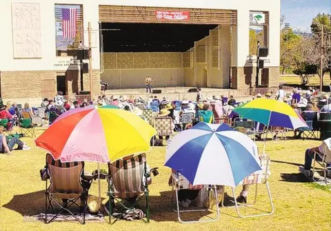 An outdoor concert venue with people seated on the lawn under colorful umbrellas watching a performer on stage.