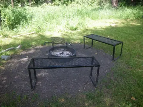 A campsite with a fire ring and two metal benches on a dirt patch surrounded by grass.