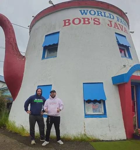Two men pose in front of a quirky building shaped like a teapot.