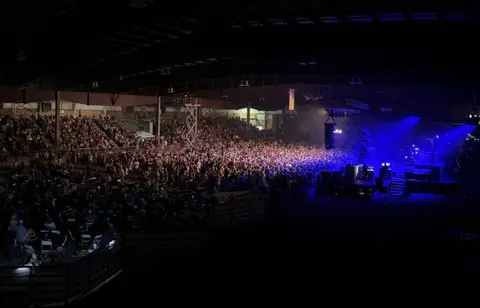 A large crowd fills an indoor arena during an event with blue stage lighting.