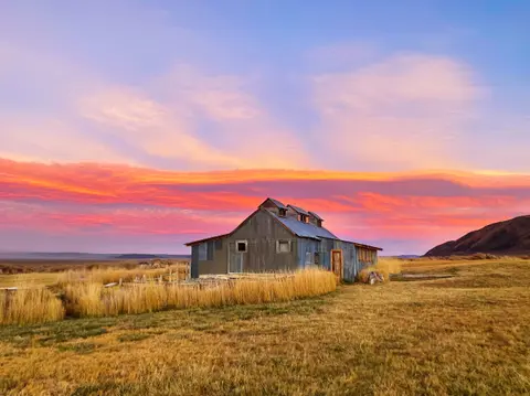 A rustic metal building stands in a dry grassy field under a colorful sunset sky.