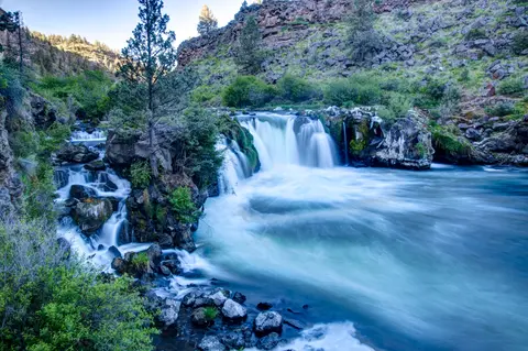 A waterfall flows into a river surrounded by rocky cliffs and trees.