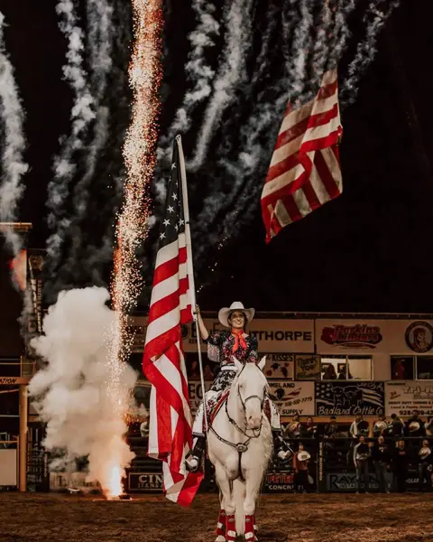 A woman on a white horse holds an American flag during an arena event with fireworks.