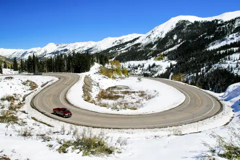 A red pickup truck drives on a winding mountain road covered in snow.