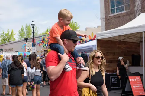 A young boy sits on a man's shoulders while walking through an outdoor event with a crowd.