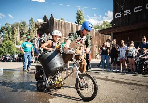 A man and child ride a custom-built wheeled contraption outside a wooden building with a crowd watching.