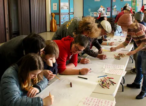 People gather around long tables to draw and write together indoors.