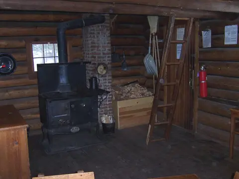 An interior view of a rustic log cabin featuring a wood-burning stove and a wooden ladder.