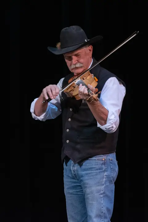 A man wearing a cowboy hat and vest plays a fiddle on stage.
