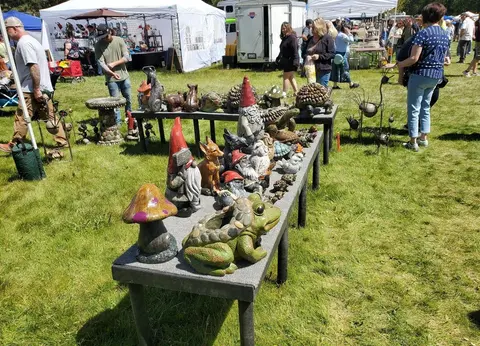 An outdoor craft market displays garden ornaments on tables in a grassy field.