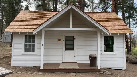 Front exterior view of a small white cabin with a wooden shingle roof and covered porch.