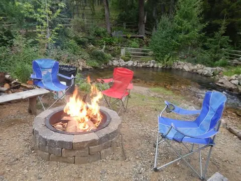 A stone fire pit with a blazing fire is surrounded by folding camping chairs near a creek and trees.