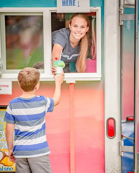 A young woman hands a snow cone to a boy through the window of a food truck.