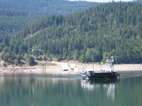 A ferry boat travels across a calm body of water with forested hills in the background.