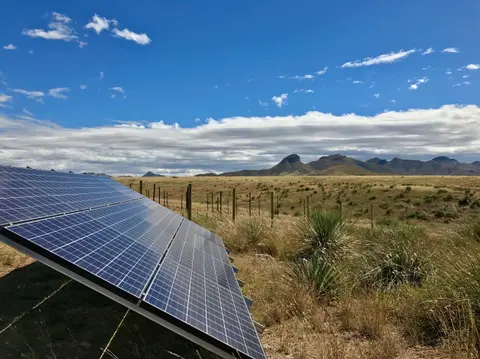 A solar panel stands in a dry field with mountains in the background under a partly cloudy sky.