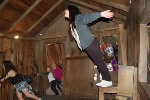 A person leans at an impossible angle inside a slanted wooden room.