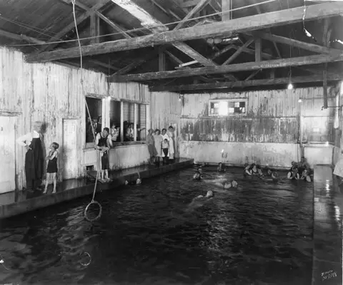 A historic black and white photograph shows people swimming in an indoor pool with a wooden structure.