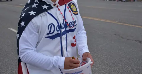 A person in a baseball jersey and an American flag cape holds a bucket outdoors.