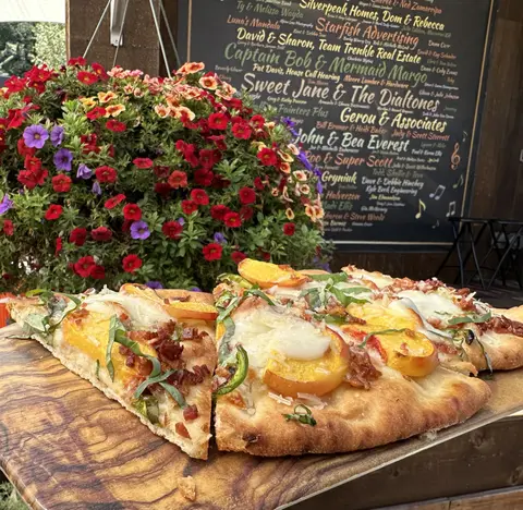 Slices of flatbread pizza sit on a wooden board next to a hanging basket of flowers.