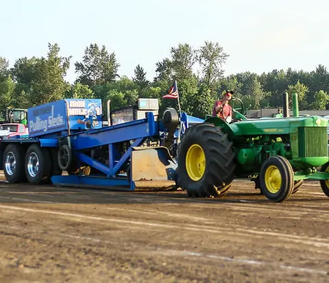 A green tractor pulls a blue sled on a dirt track.