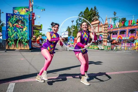 Two performers in colorful costumes hold a hula hoop outdoors at a fairgrounds.