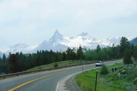 A car drives along a winding road with snowy mountains in the background.