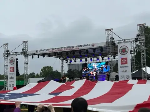 A large American flag is held up in front of an outdoor festival stage.