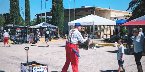 A street performer in red pants and a blue hat performs at an outdoor market.
