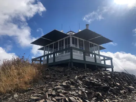 A fire lookout cabin sits on a rocky mountain peak under a blue sky with clouds.