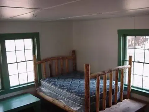 A rustic wooden bed frame with a mattress is situated in a small room with two windows.