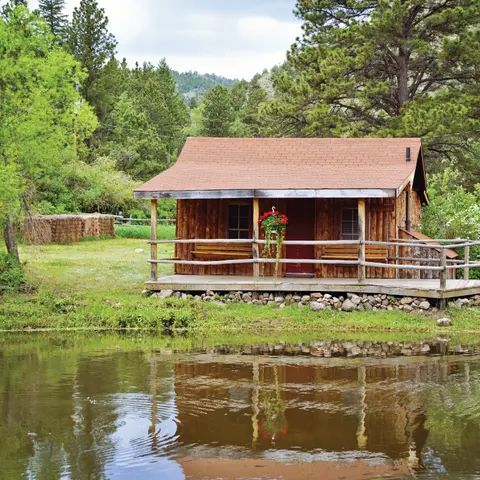 A rustic wooden cabin with a porch sits beside a body of water, surrounded by trees and greenery.