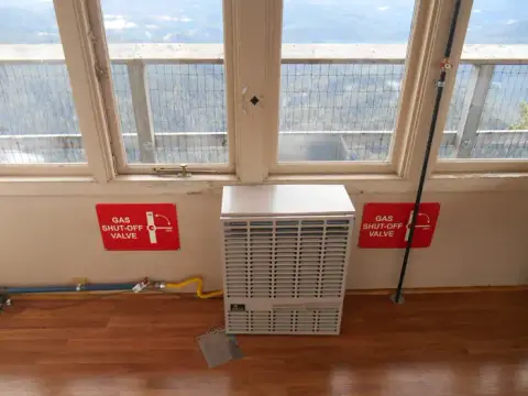 An indoor view of a wall heater under windows with red gas shut-off valve signs.