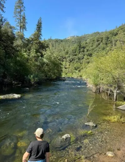 A person looks out over a river surrounded by trees and mountains.